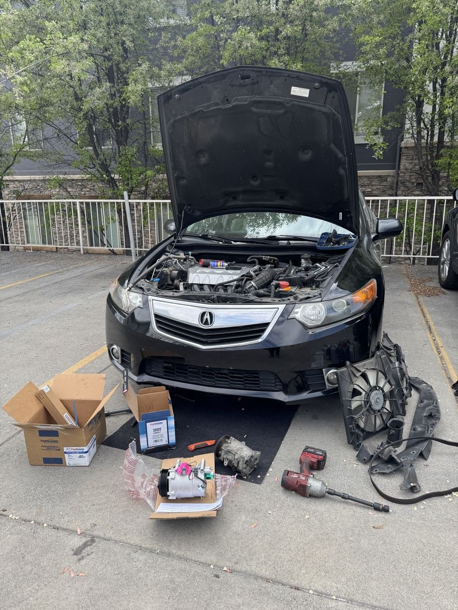 Acura TSX with the hood up and a new AC compressor and parts boxes on the ground