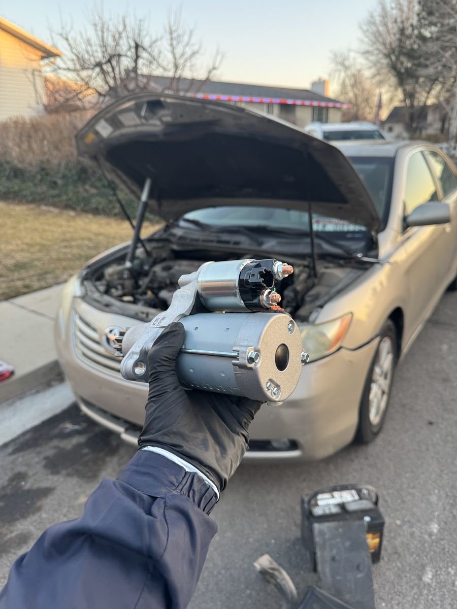 Holding a new starter in front of a Toyota Camry with the hood up